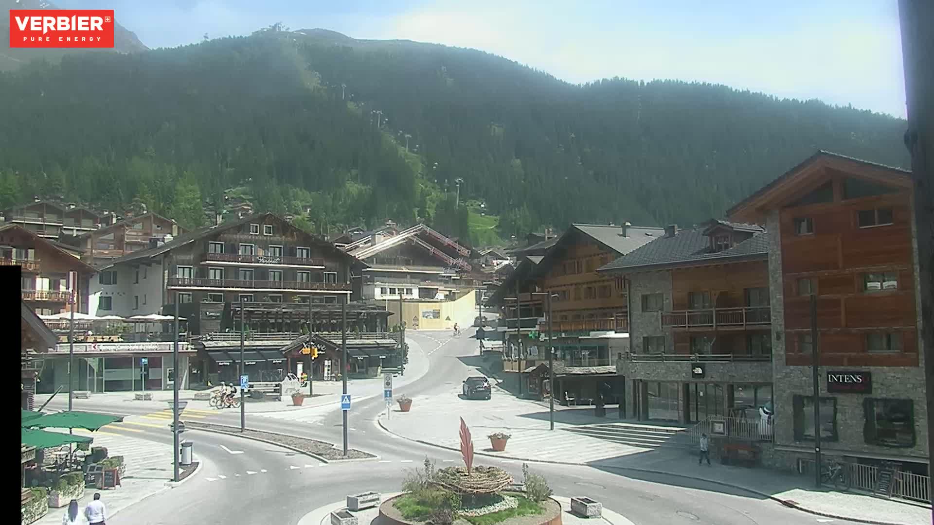 A sunny mountain village square with several multi-story buildings, a roundabout, and a few people walking around.