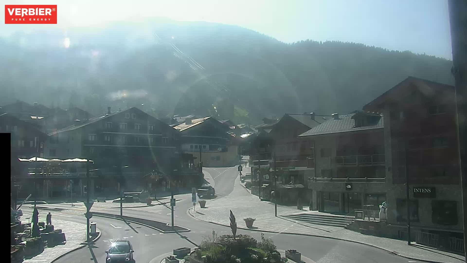 A hazy, sunny day reveals a mountain village square with several buildings, a few cars, and people walking around.