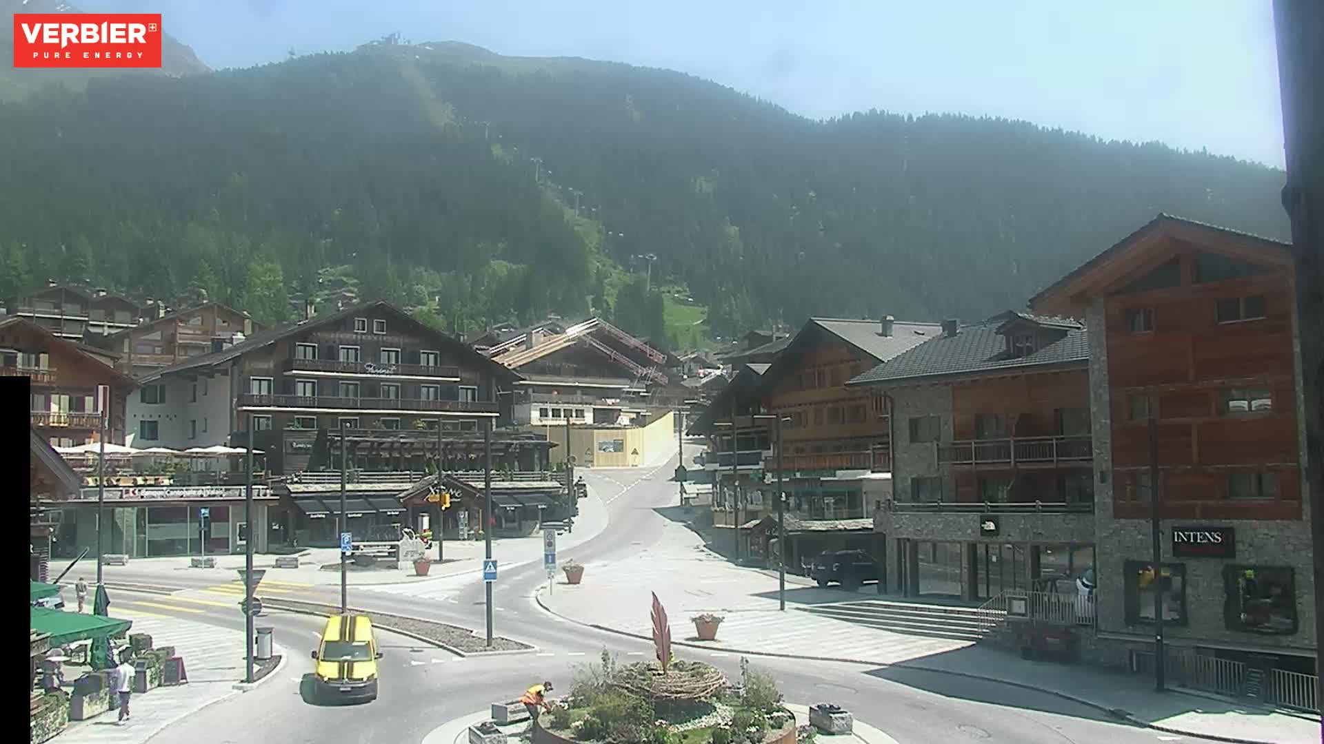 A sunny view of a town nestled in a valley, with mountains in the background and several buildings lining a paved square.