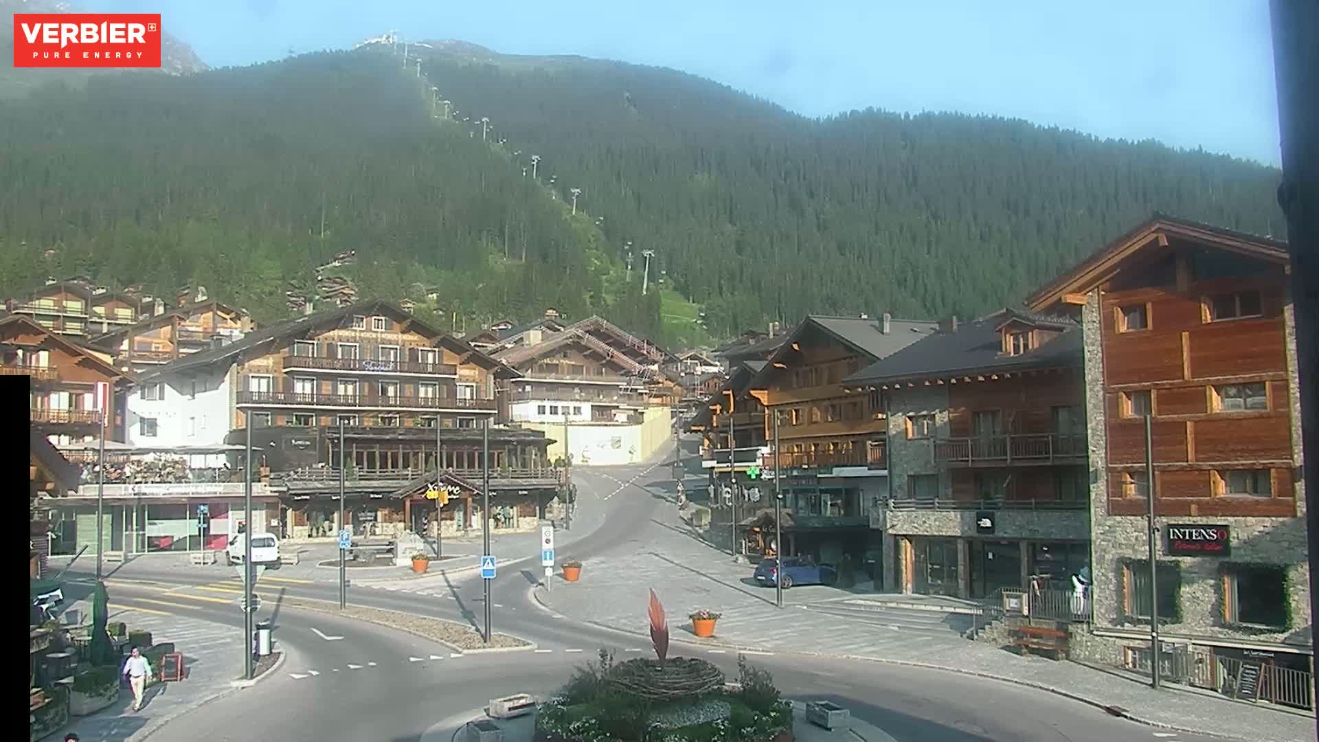 A mountain village square with chalet-style buildings is seen on a clear, sunny day, with a forested mountain rising behind it.