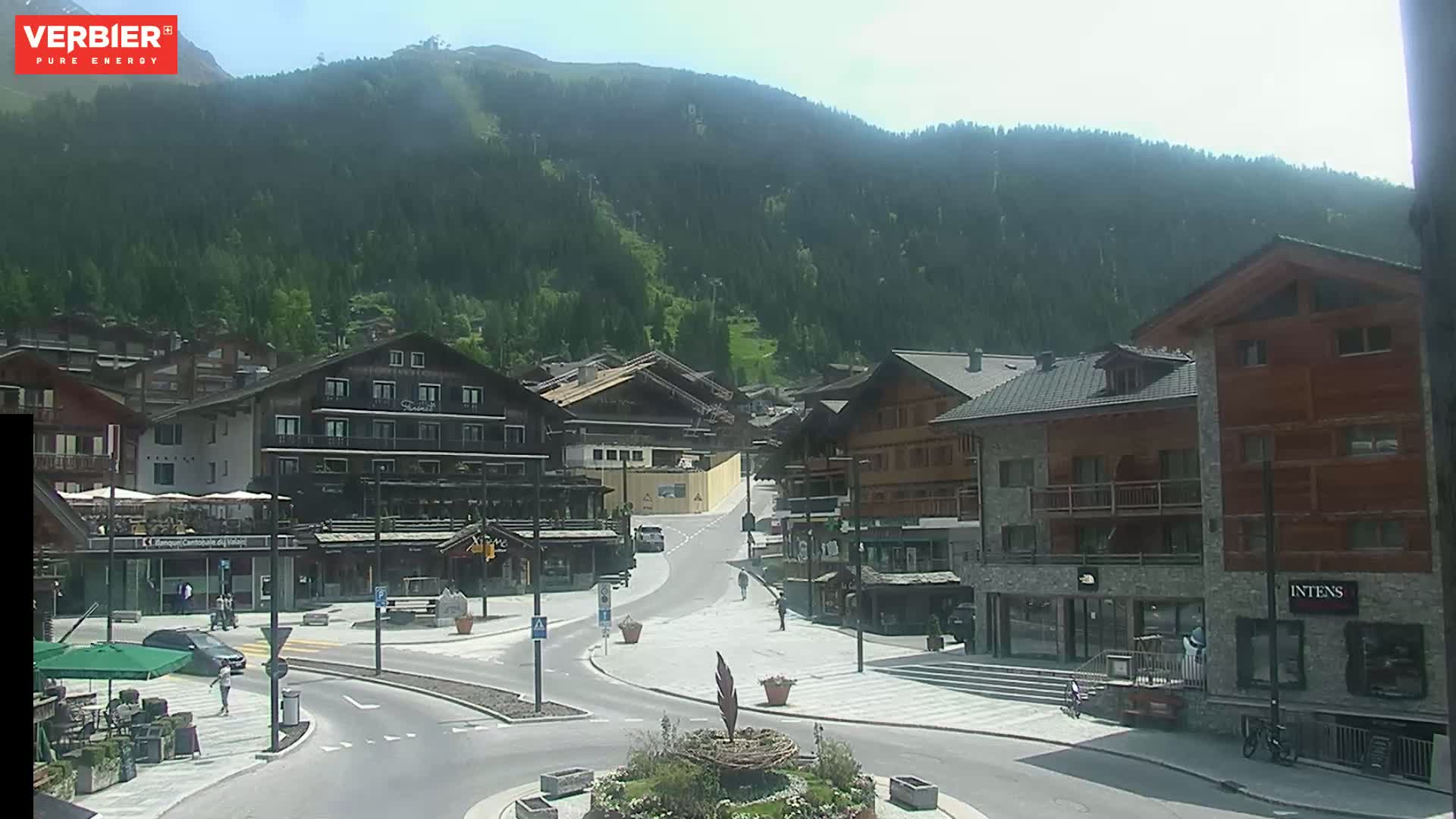 A sunny view of a village nestled in a mountainous region, with several buildings lining a paved road that leads towards the forested slopes.