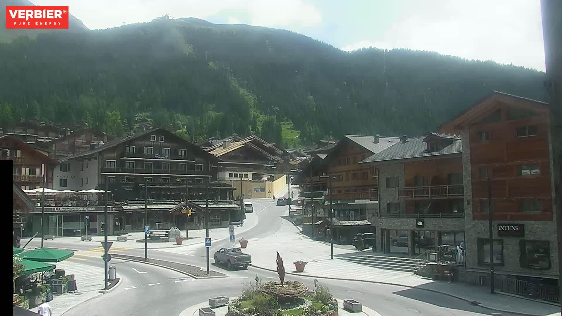 A sunny day in a mountain village with several buildings surrounding a town square, a pickup truck driving through the area, and a forested mountain in the background.