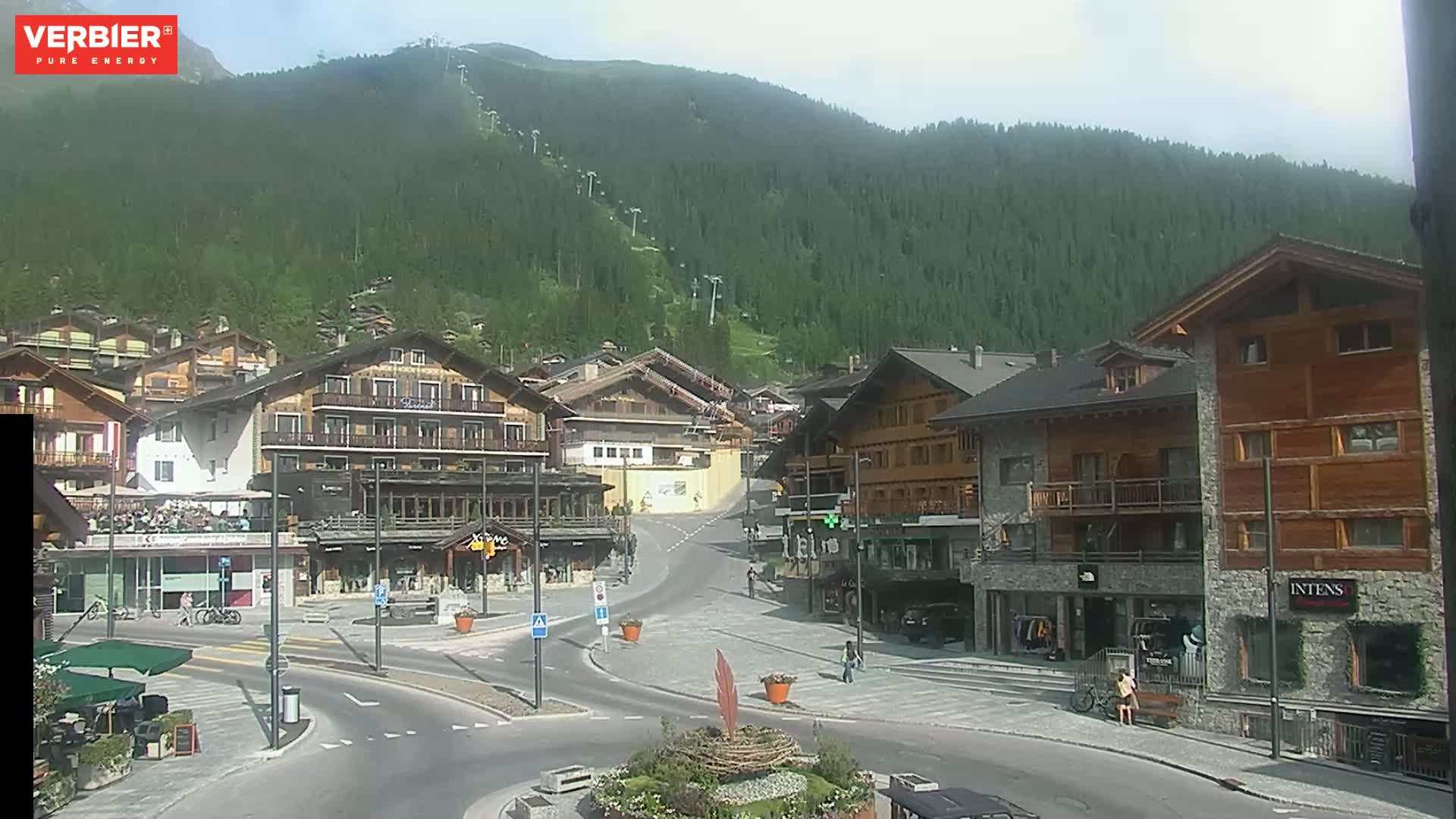 A mostly sunny view of a town nestled in a valley, surrounded by a lush green mountainside with ski lifts visible.