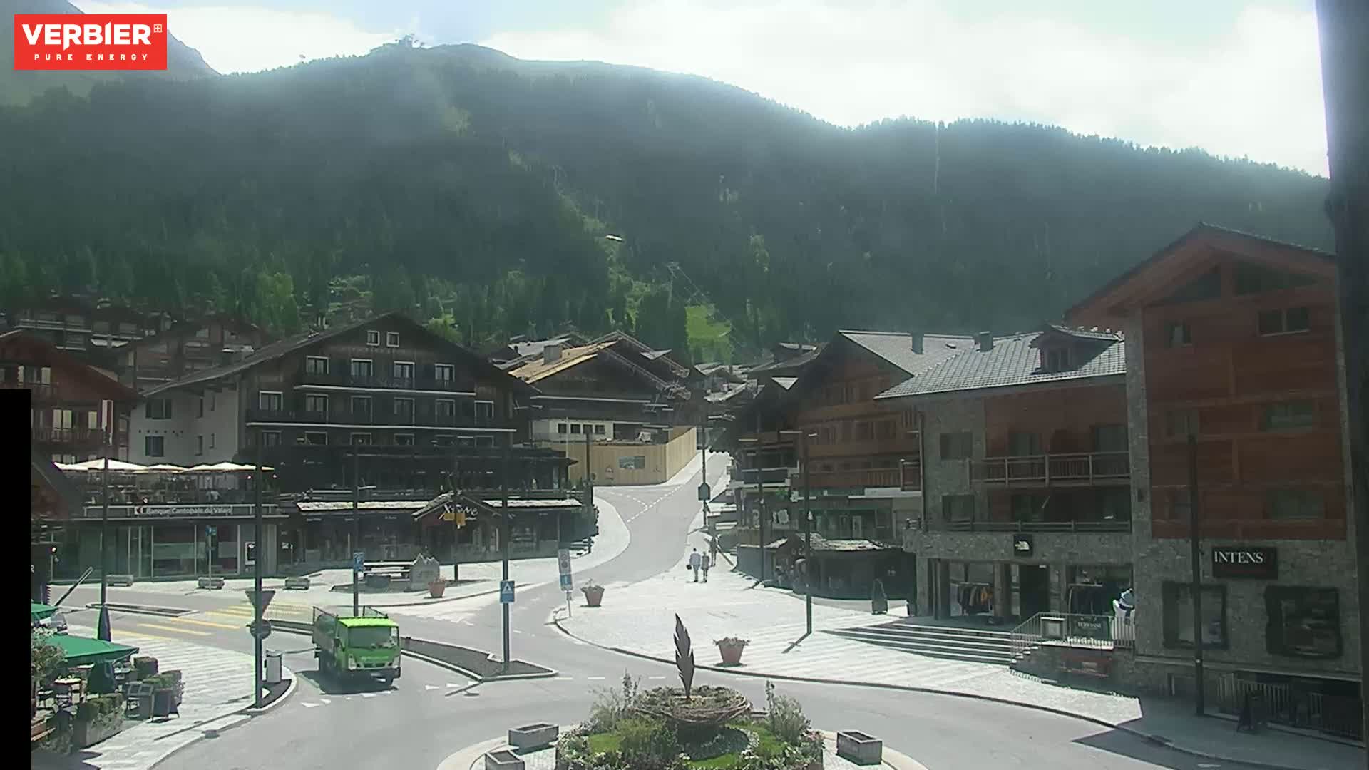 A sunny mountain village square with several buildings, a small roundabout, and a few people walking.