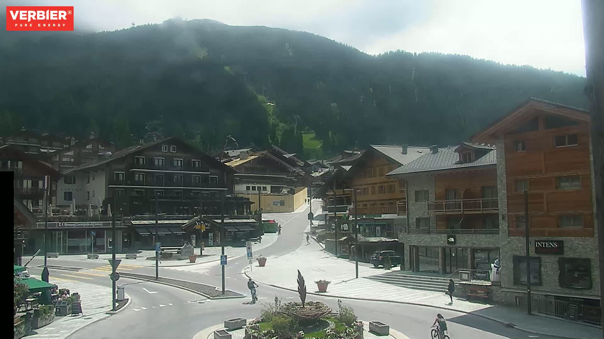 A sunny day in a mountain village shows a plaza with several buildings, a roundabout, and a few pedestrians and cyclists.