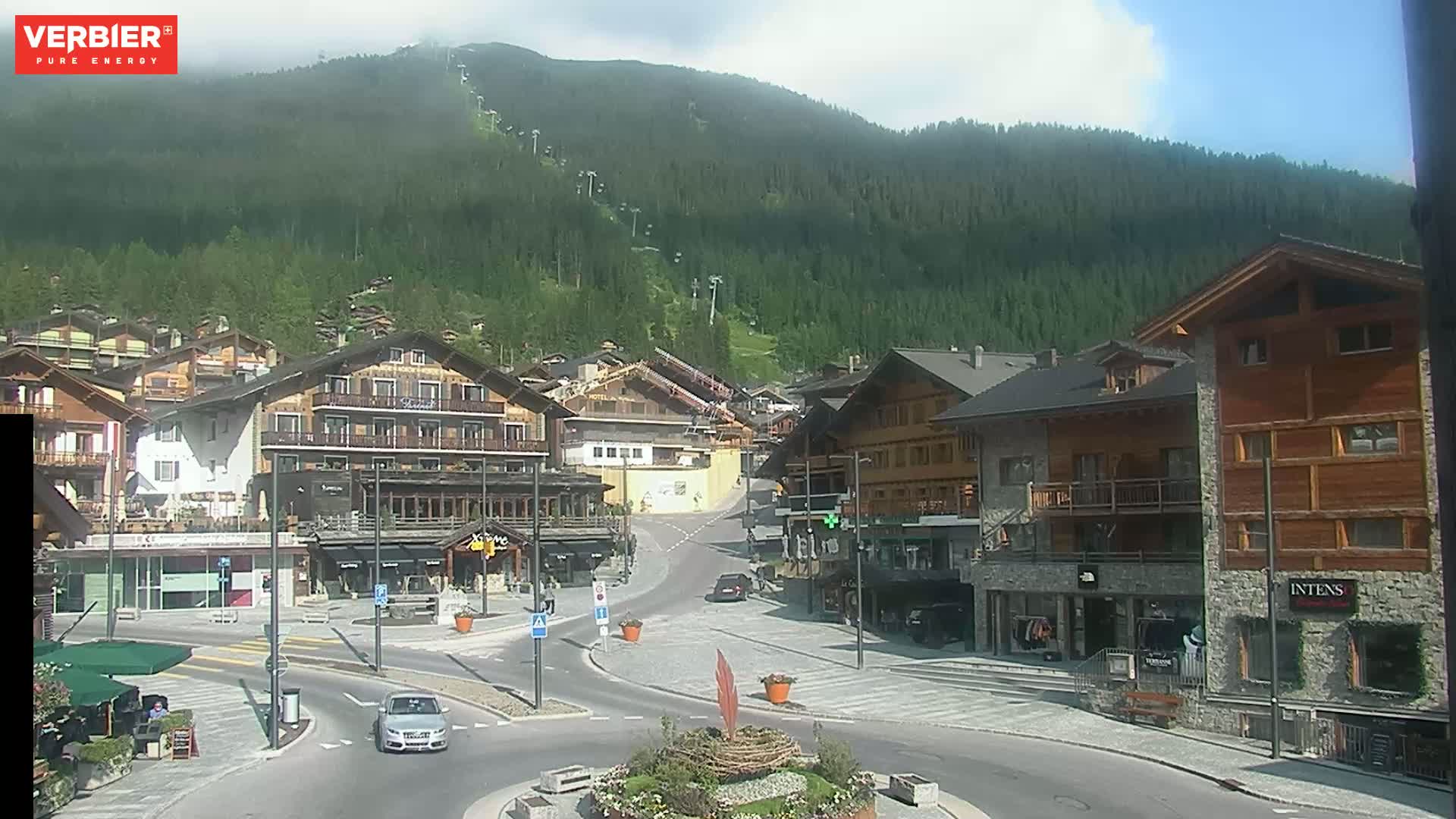A mostly sunny view of a village nestled in a valley with a mountain covered in green trees in the background.