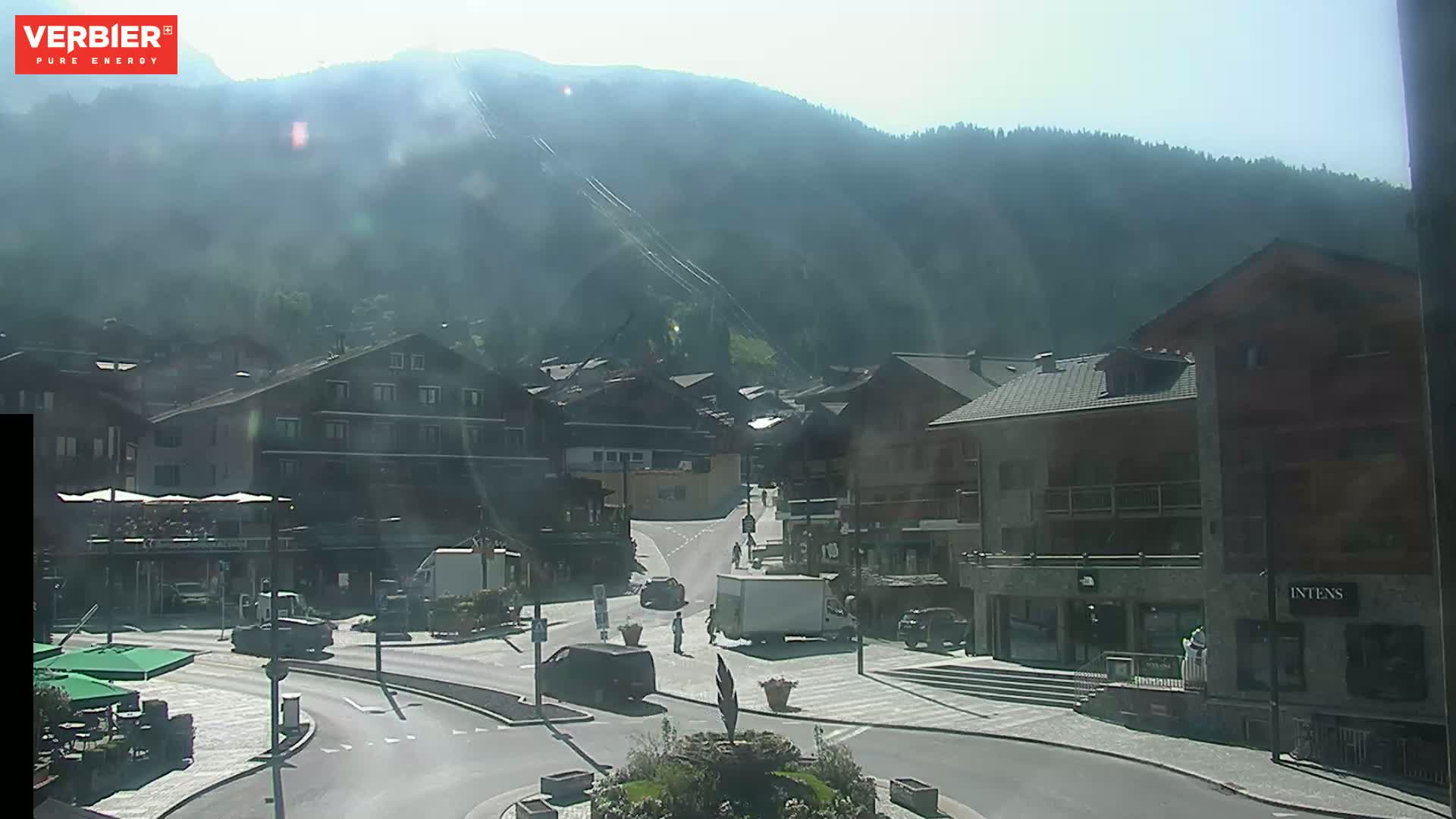 A sunny day in a mountain village with several buildings and vehicles in a town square, backed by a hazy mountain range.