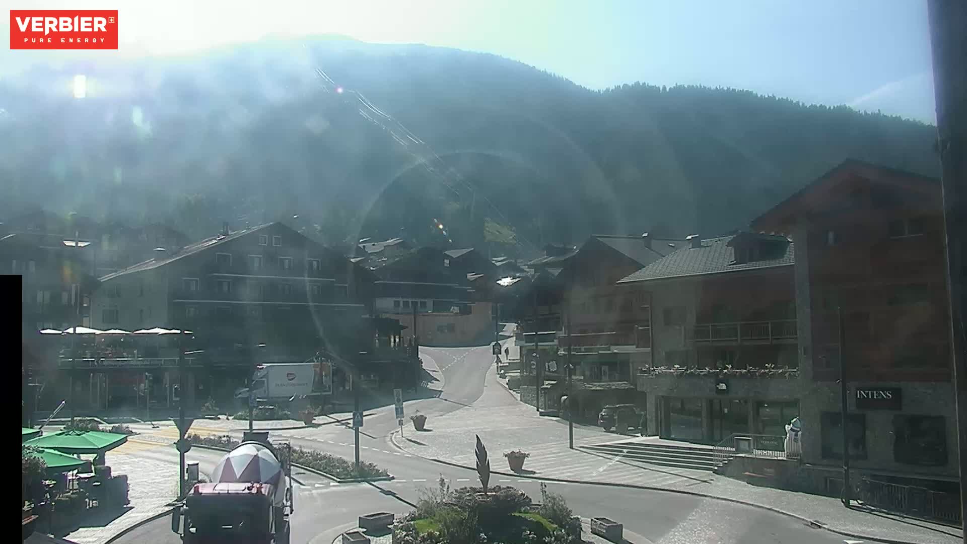 A hazy, sunny day reveals a mountain village square with buildings, a cement mixer, and a roundabout, all situated against a backdrop of a mountain range.