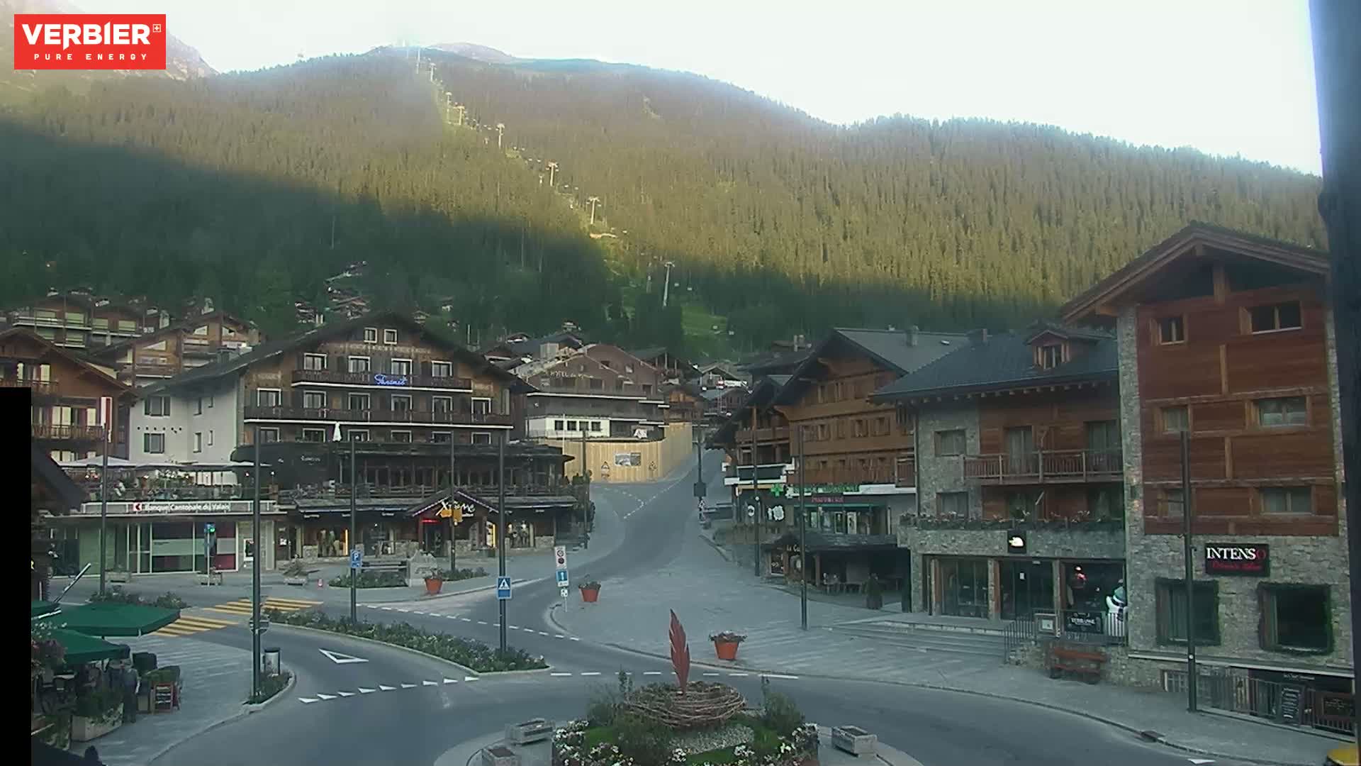 A mostly empty town square in a mountain village on a partly sunny day, with a forested mountain rising behind it.