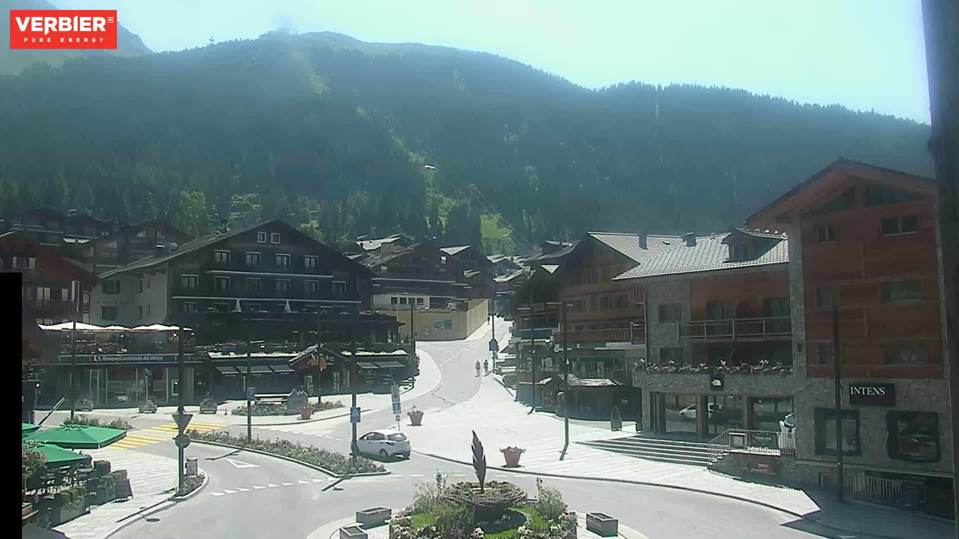 A sunny day in a mountain village shows a plaza with buildings, cars, and people walking on a road leading towards a forested hillside.
