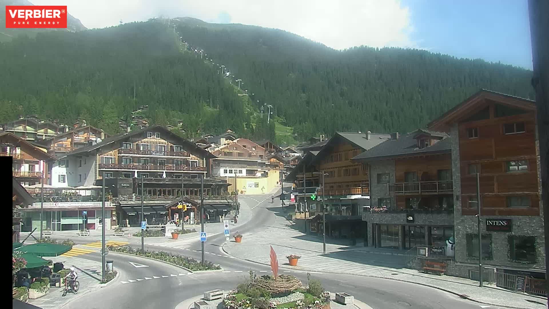 A mountain village square with several multi-story buildings, a ski lift ascending a forested mountain in the background, under a mostly sunny sky.