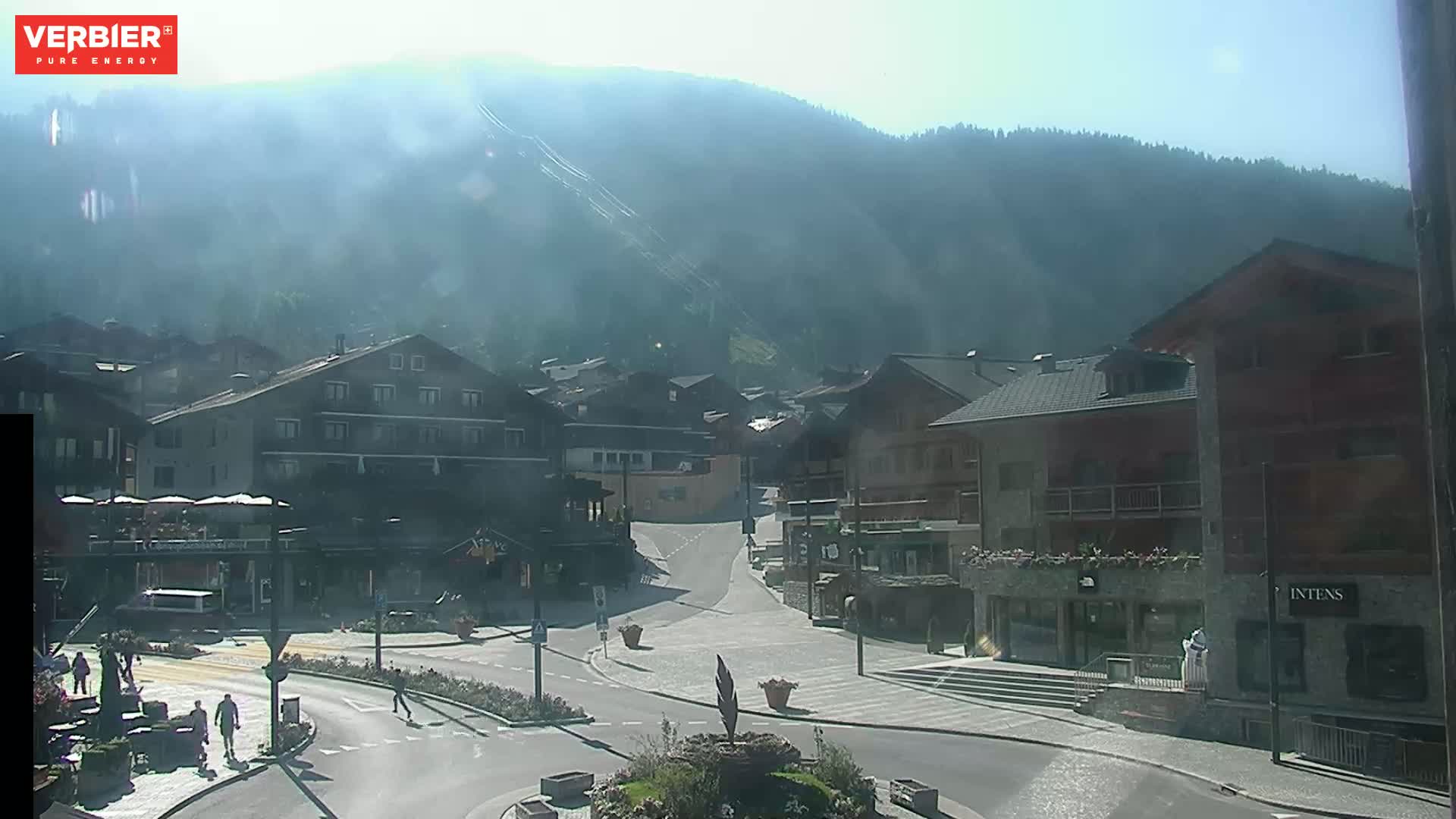 A hazy, sunny day reveals a mountain village square with several buildings, a roadway, and a few pedestrians.