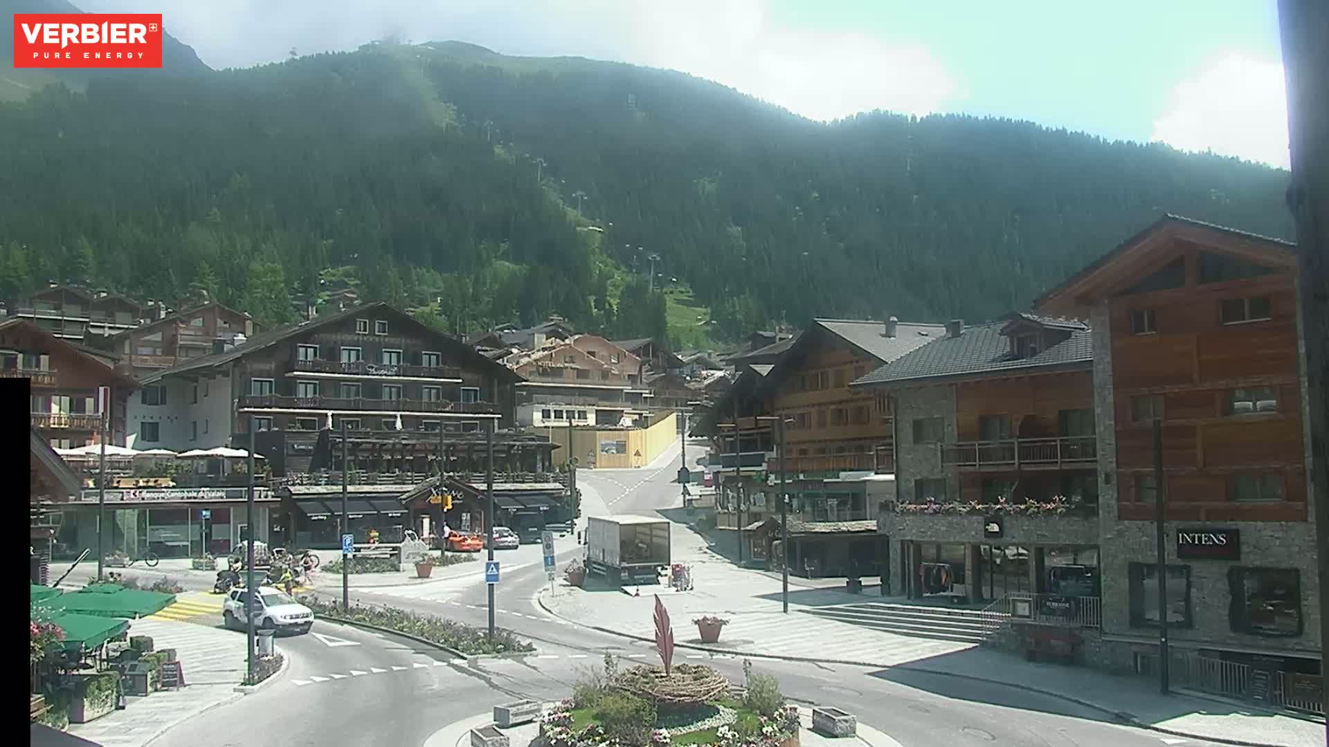 A sunny day in a mountain village with several buildings surrounding a plaza, a few cars parked, and a mountain covered in green trees in the background.