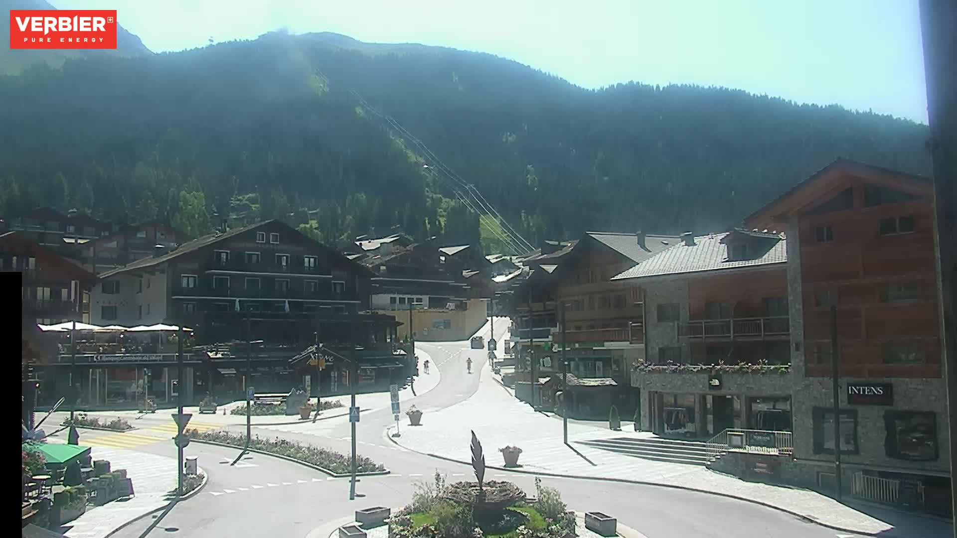 A mostly sunny view of a village nestled in a mountain valley, with several buildings lining a paved street.