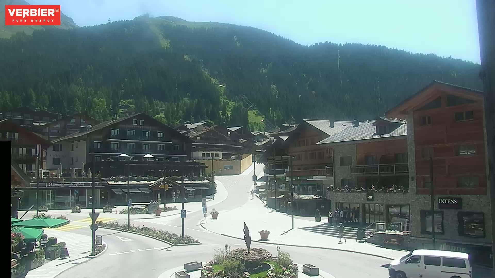 A sunny mountain village square with several buildings, a roundabout, and a few people.