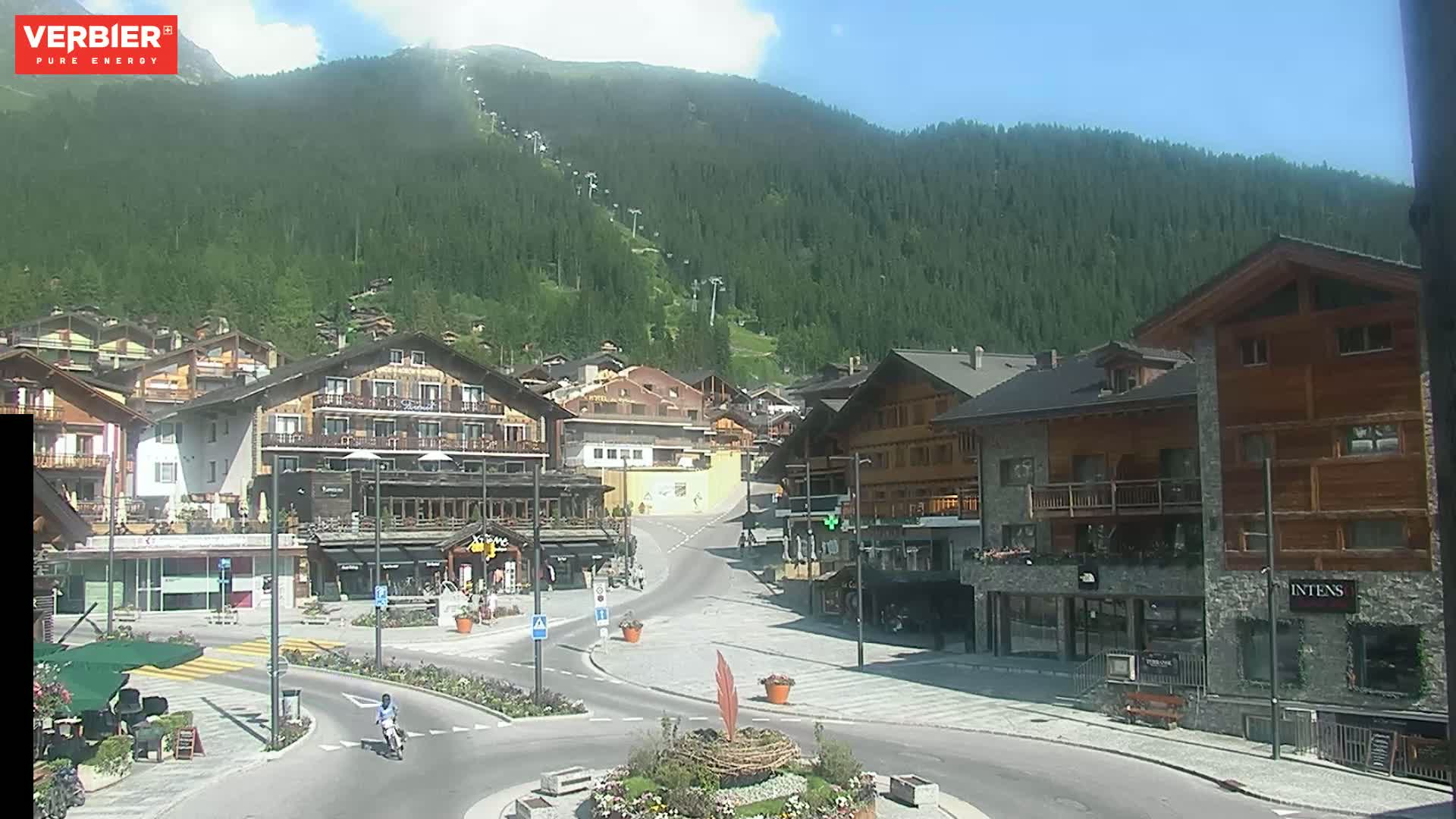 A mountain village square with several multi-story buildings, a person on a scooter, and a mostly sunny sky.