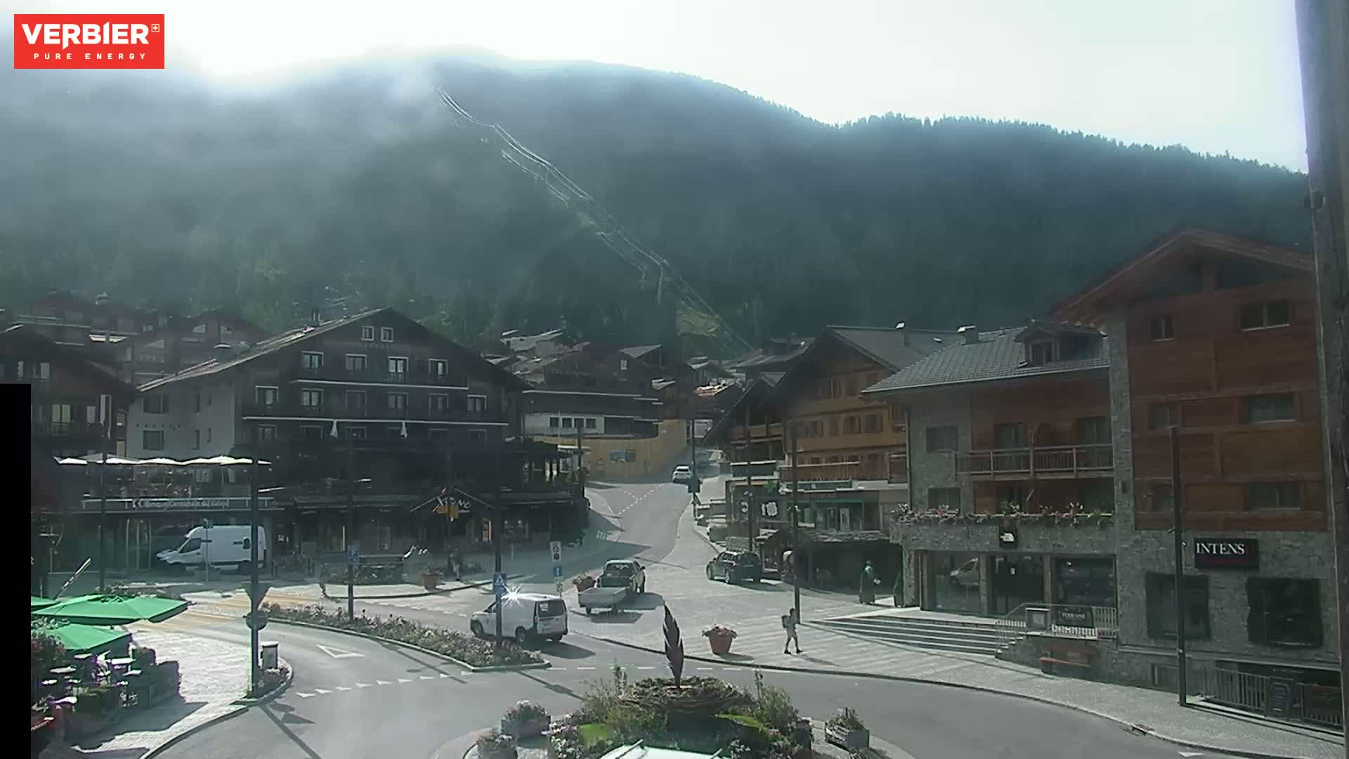 A hazy, sunny day reveals a mountain village scene with several buildings, a roadway, and a few vehicles, all nestled against a backdrop of a mountain covered in trees.