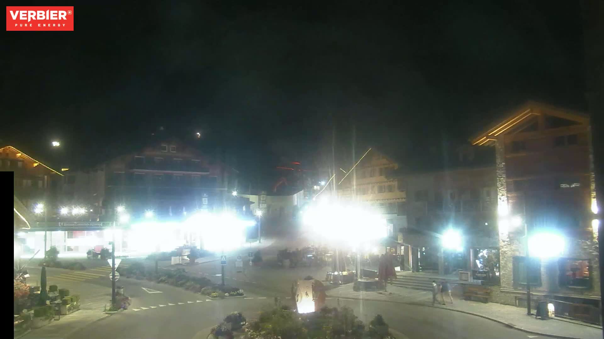 A nighttime view of a town square with several buildings illuminated, under a clear dark sky.