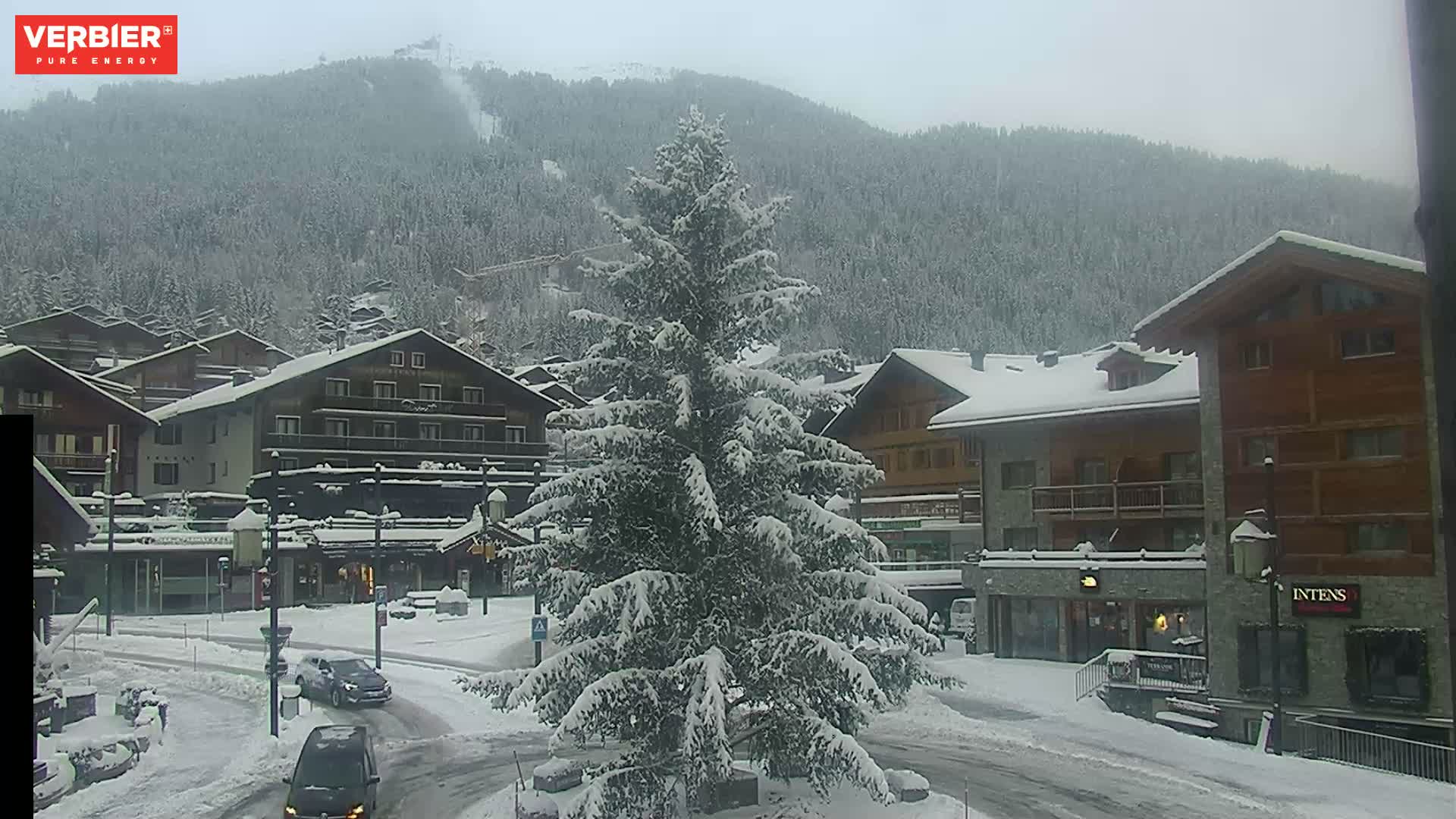 A mountain village is blanketed in snow under an overcast sky, featuring traditional chalet-style buildings, a large snow-laden evergreen tree, and two cars driving on a snow-covered road.