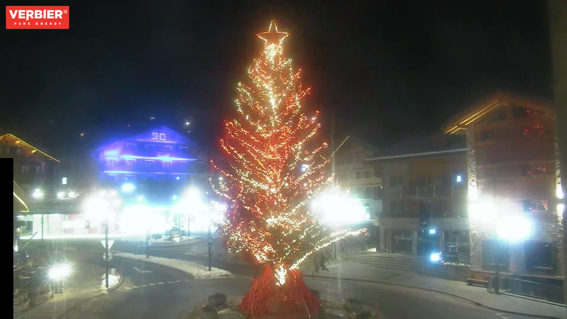 A large, star-topped Christmas tree adorned with warm lights illuminates a snowy village intersection at night, surrounded by buildings and streetlights under clear skies.