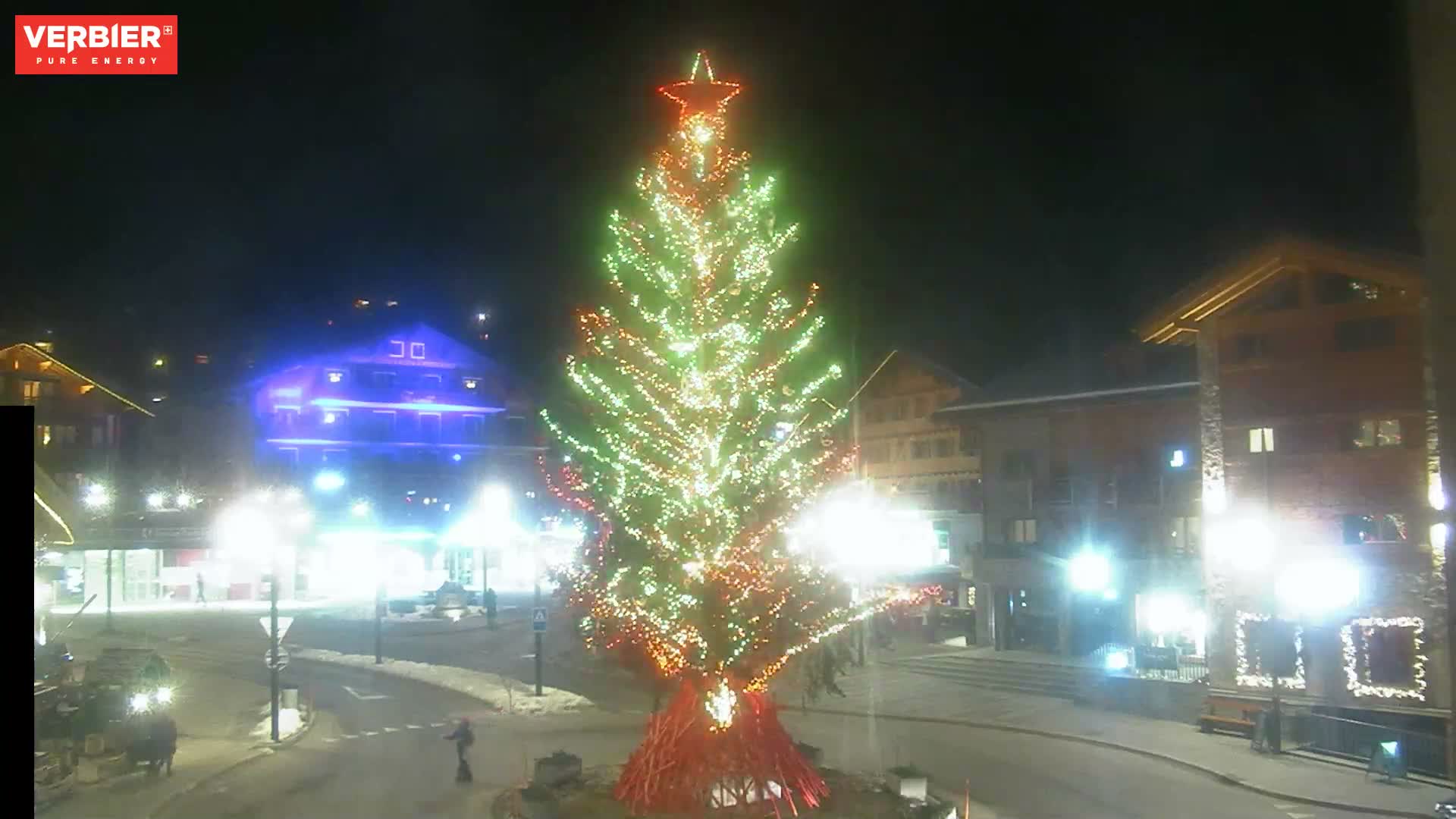 A large, star-topped Christmas tree adorned with warm lights illuminates a snowy village intersection at night, surrounded by buildings and streetlights under clear skies.