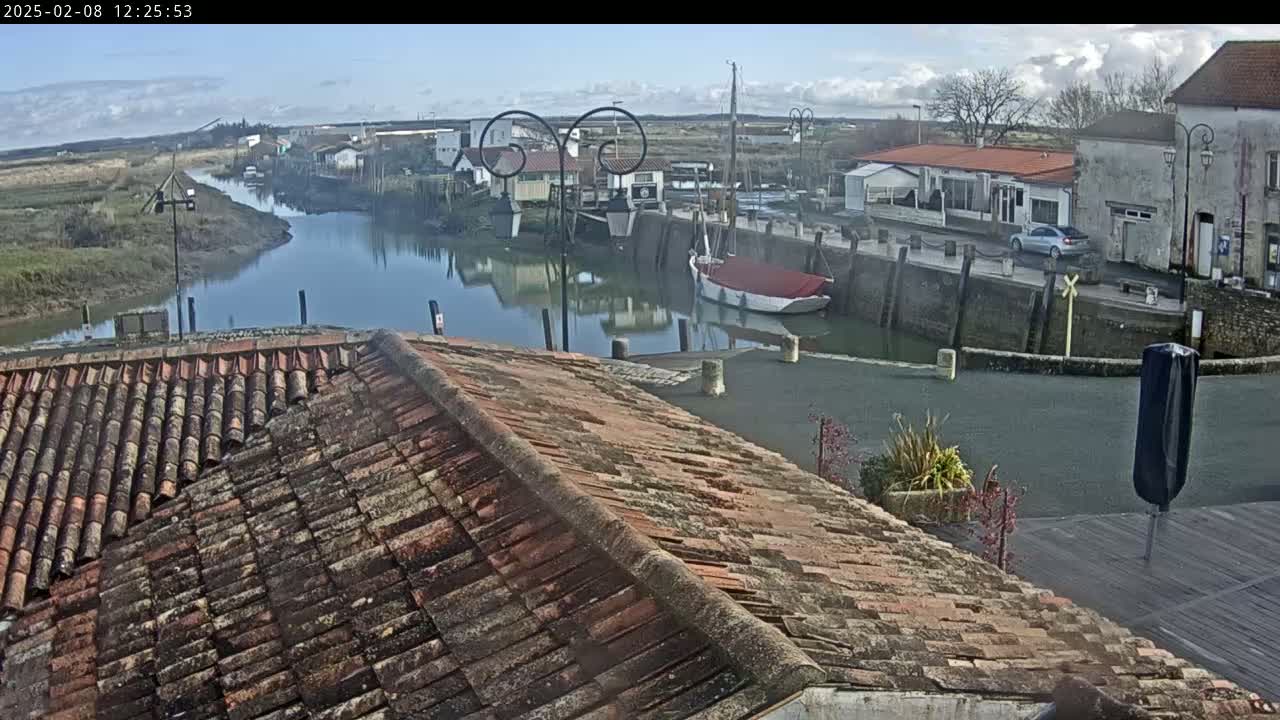 A mostly sunny day overlooks a calm river that winds through a small village with docked boats and buildings with terracotta roofs.