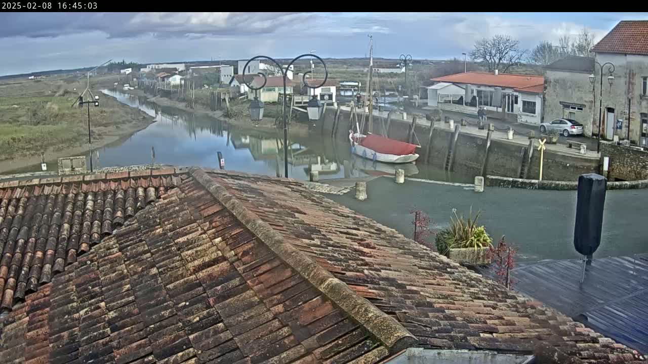 A red-roofed building overlooks a calm waterway, where a sailboat is moored near a small village under a cloudy sky.