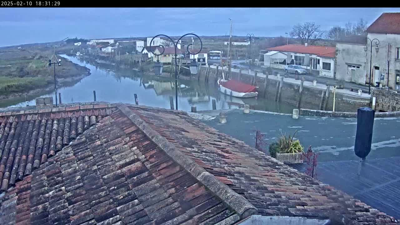 A view from a tiled roof shows a calm waterway with docked boats and buildings along its banks under an overcast sky.