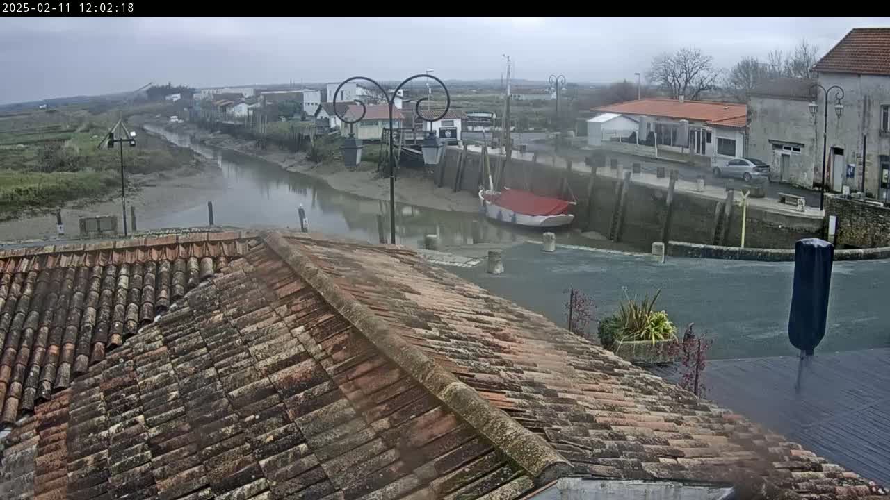 A view from a tiled roof overlooks a calm waterway in a small town on an overcast day, with a sailboat docked near buildings and a car parked nearby.