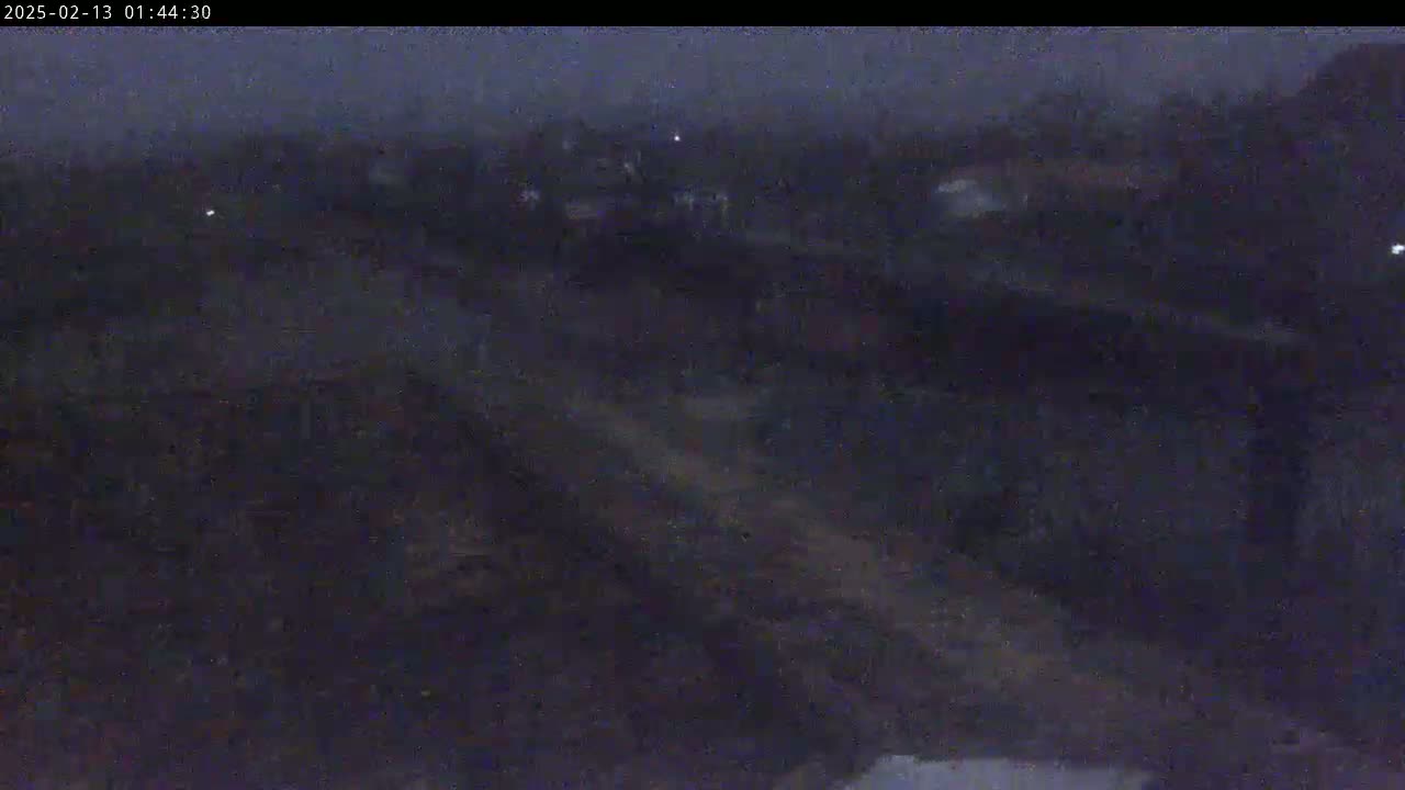 A dark, nighttime scene shows a mostly obscured landscape with faint lights in the distance under a cloudy sky.