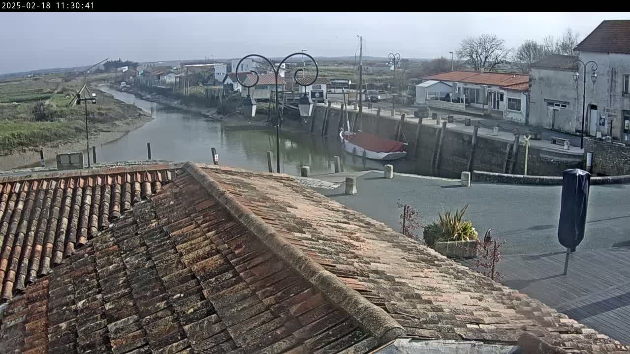 A view from a tiled roof overlooks a calm waterway, a small harbor with boats, and a village of buildings under a clear, sunny sky.