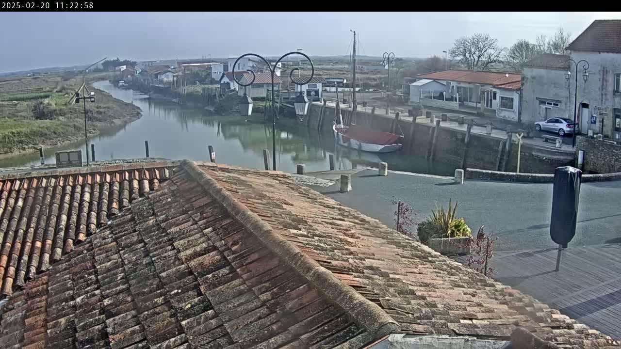 A view from a rooftop shows a calm waterway winding through a small village on a sunny day, with boats moored along the quay.