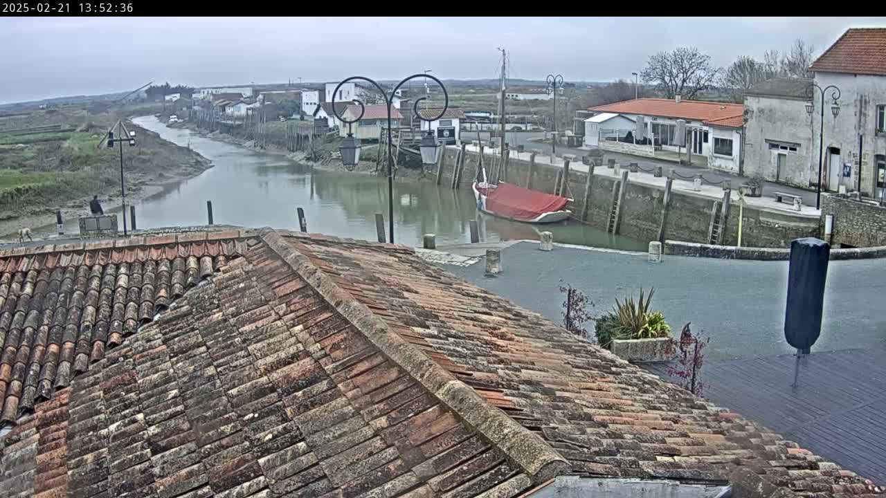 A red-sailed boat is moored in a calm harbor next to a village on an overcast day, viewed from a tiled roof.