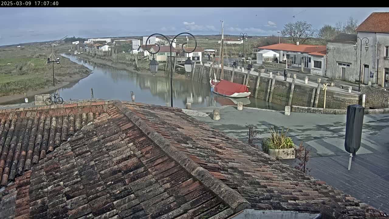 A mostly sunny day overlooks a calm waterway next to a village with a red-covered boat docked, viewed from a tiled roof.