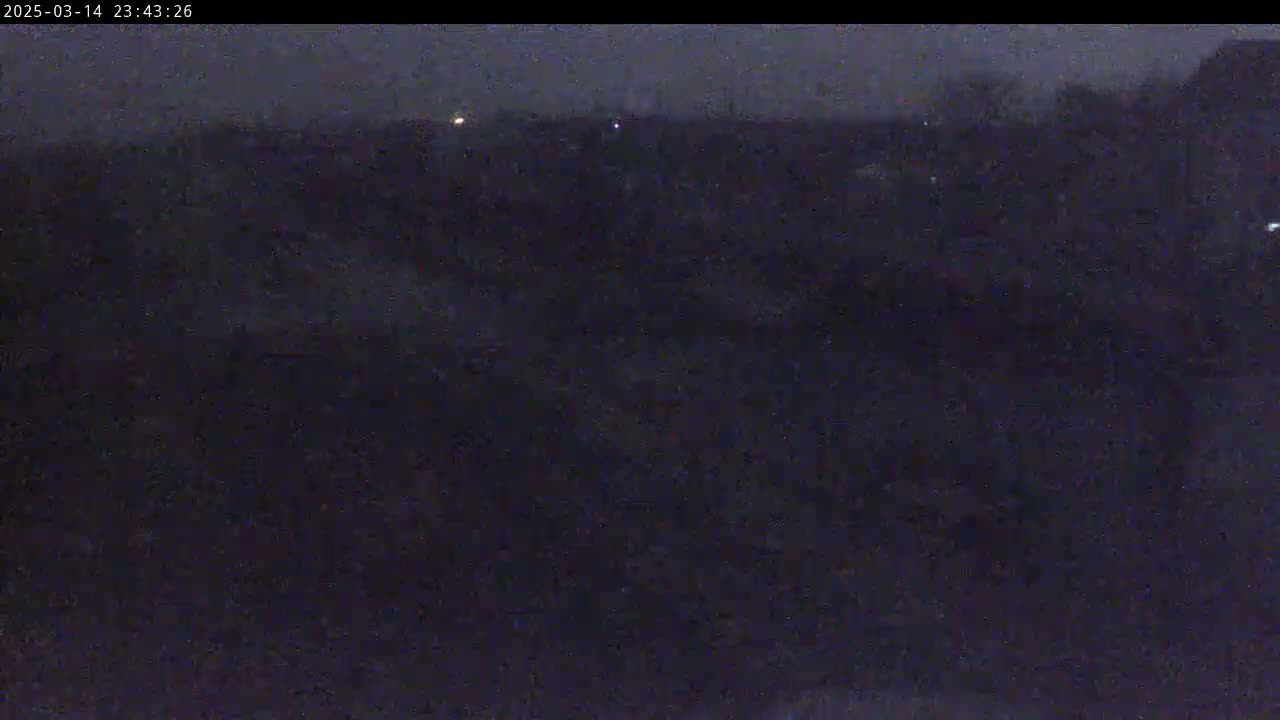 A dark, nighttime outdoor scene shows a mostly obscured landscape with a few faint lights visible in the distance under a possibly snowy or hazy sky.