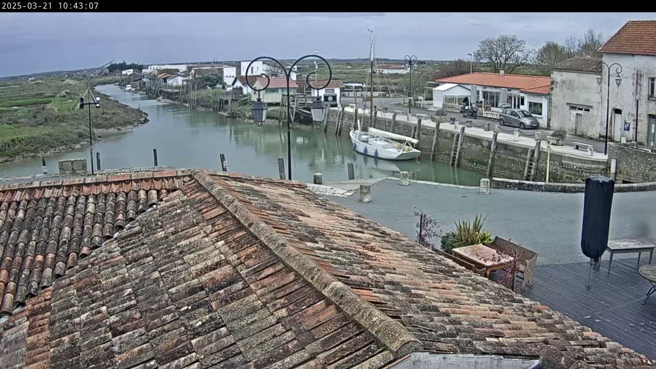 A view from a tiled roof overlooks a calm waterway with docked boats and buildings along the shore under an overcast sky.