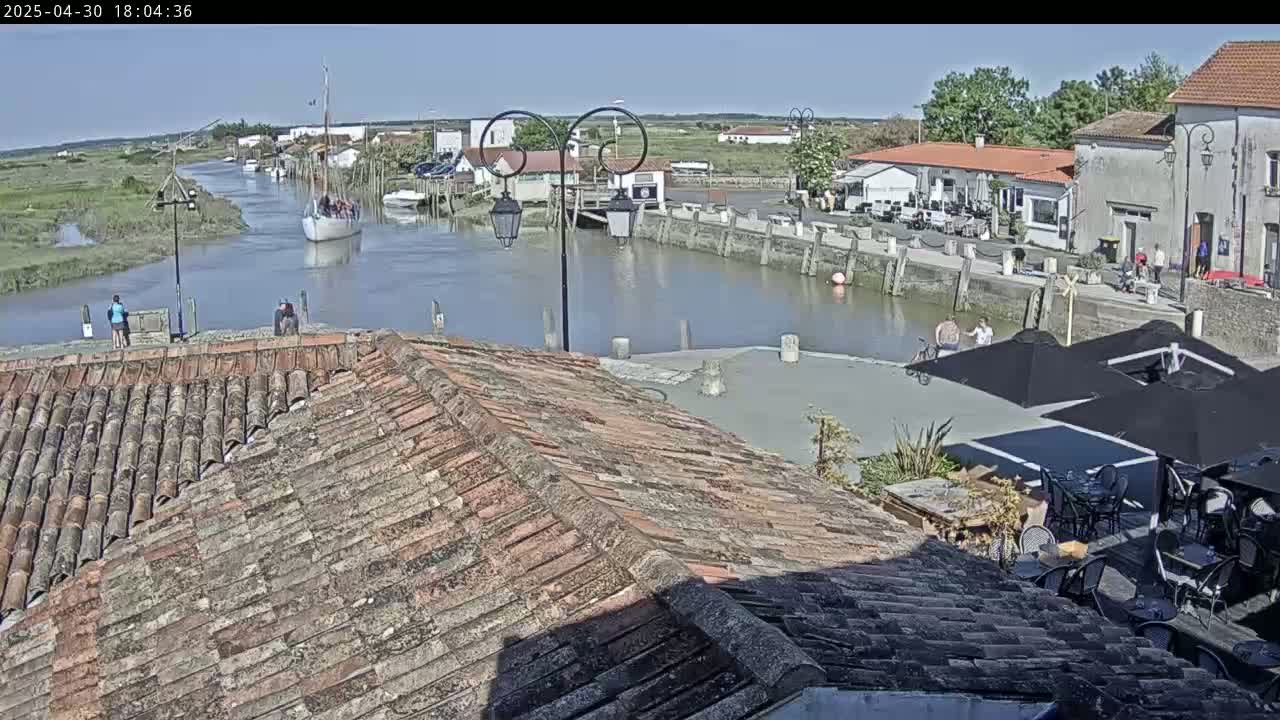A sailboat is in a calm waterway next to a village with a tiled roof overlooking an outdoor patio area under sunny skies.