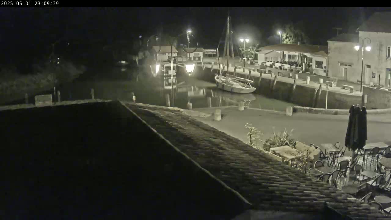 A nighttime view of a small harbor with a sailboat, buildings, and outdoor seating, under clear skies.