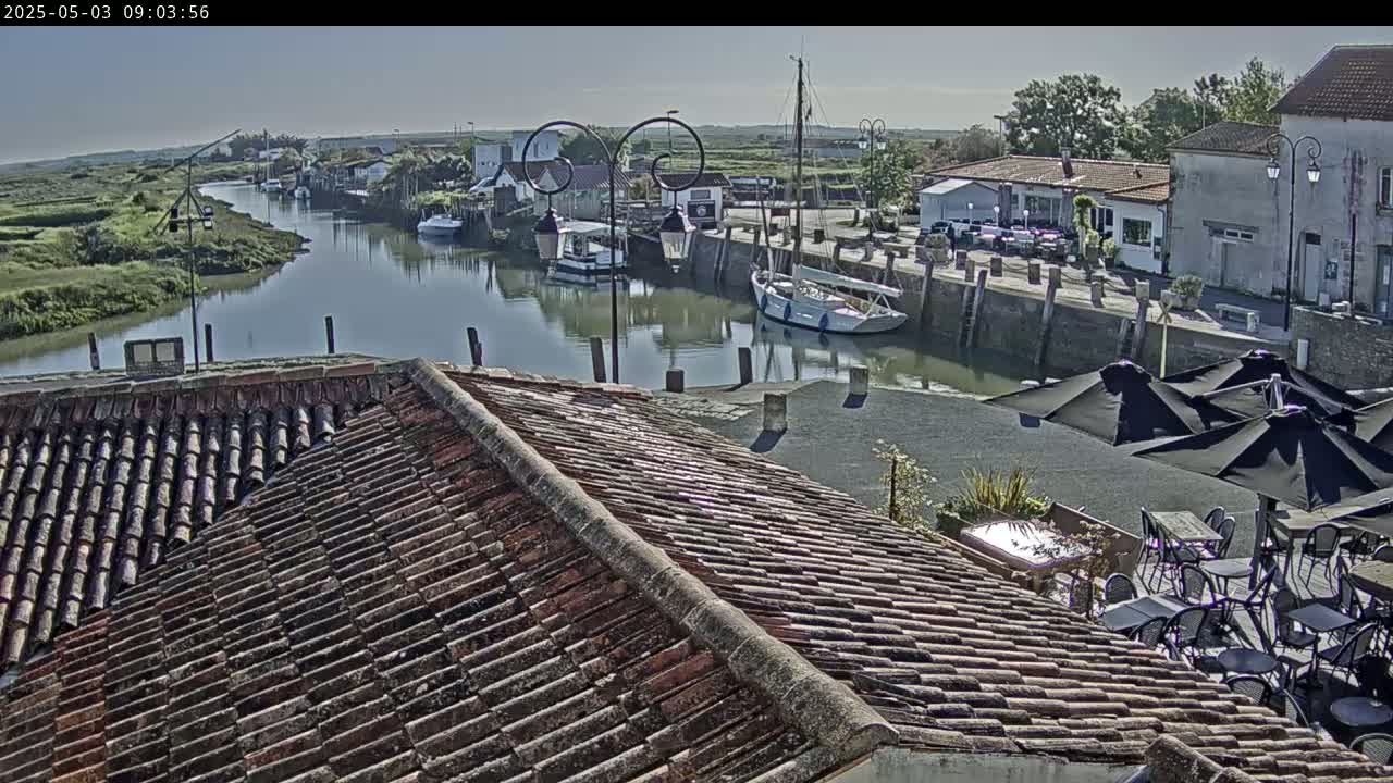 A tile roof overlooks a calm waterway with moored boats and a waterfront village under a clear sky.