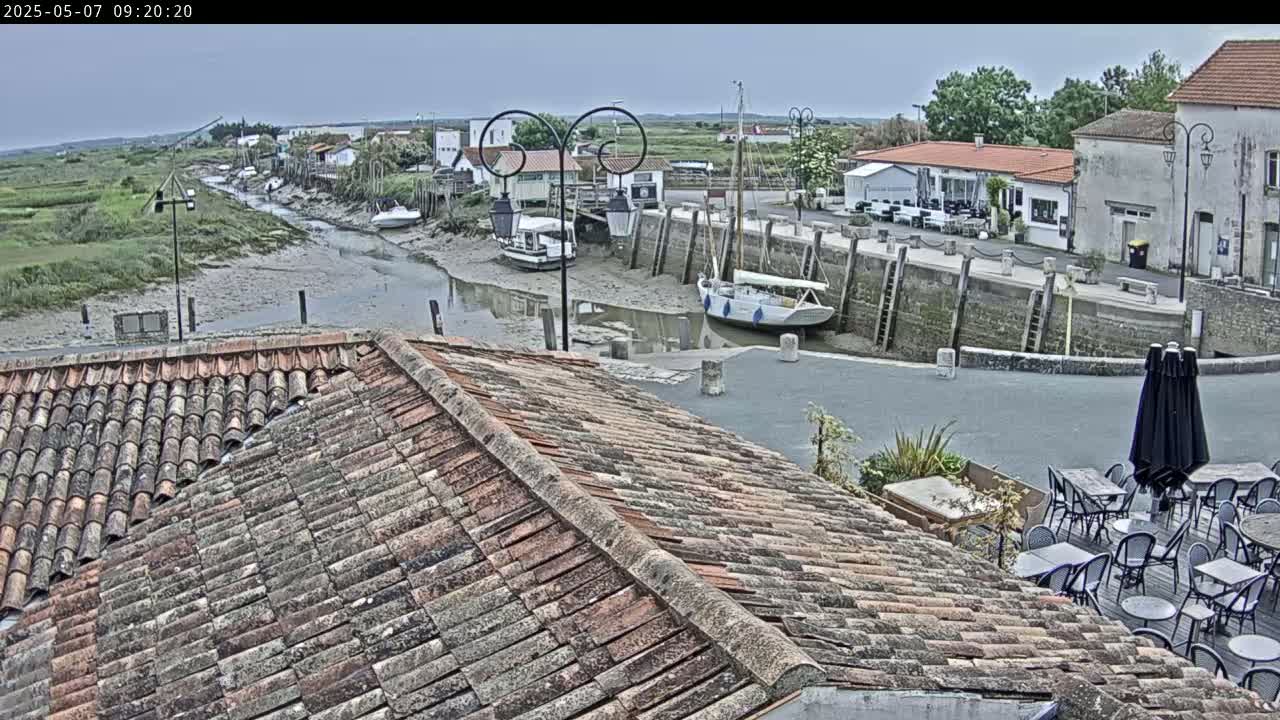 A view from a tiled roof shows a small harbor with boats at low tide, under an overcast sky, and an outdoor cafe with tables and chairs.