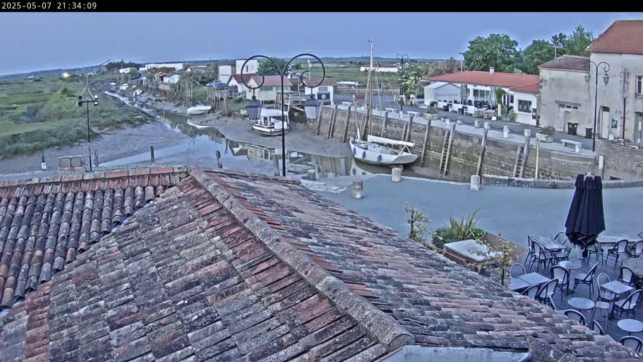 A tiled roof overlooks a calm harbor at low tide, with several boats moored near buildings under a twilight sky.