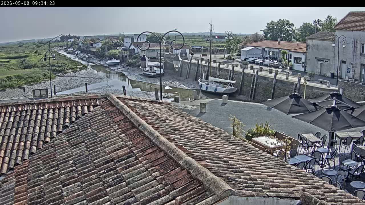 A tiled roof overlooks a calm harbor scene on a sunny day, with several boats docked and a patio with umbrellas and tables below.