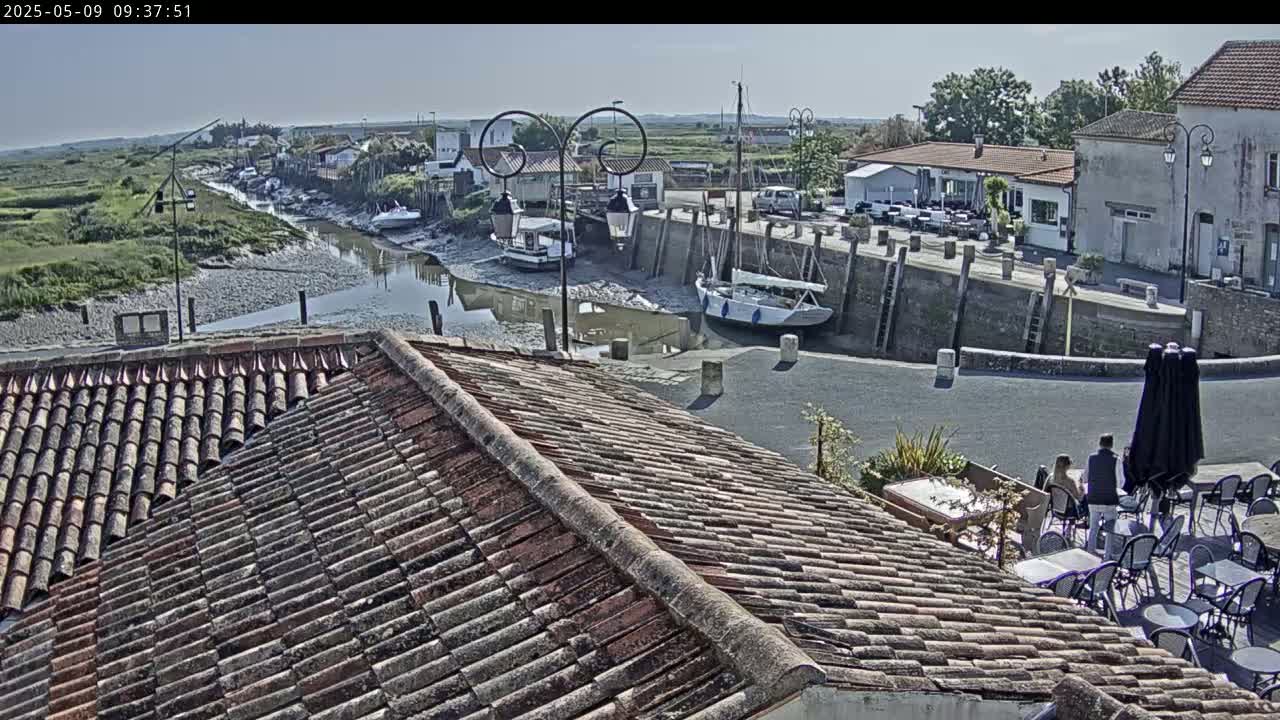 A tiled roof overlooks a small harbor with boats at low tide on a sunny day, with a few people sitting at outdoor tables near a building.