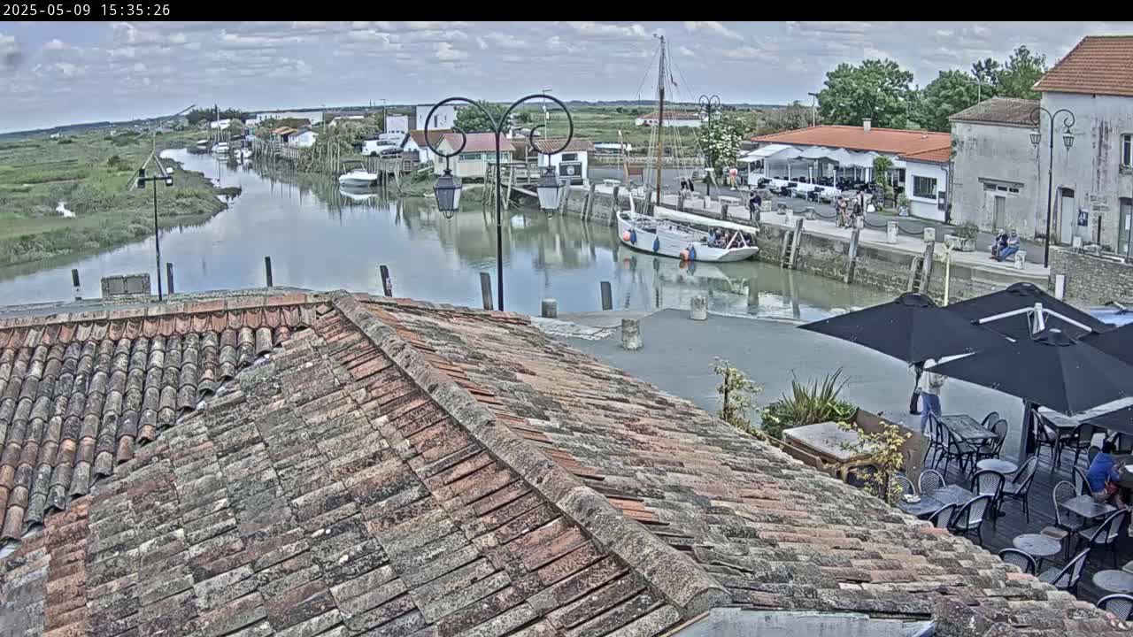 Overlooking a calm waterway, a tiled roof provides a view of a quaint village with boats moored along a canal under a partly cloudy sky.