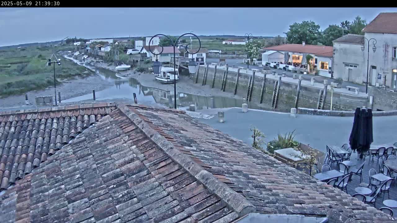 A tiled roof overlooks a calm, shallow waterway at dusk, with a small village and outdoor seating area visible in the background.