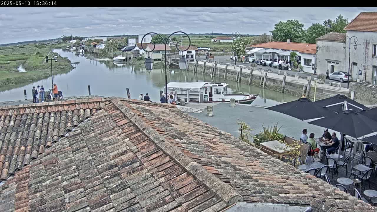 A tiled roof overlooks a waterway where a small boat is docked near buildings with outdoor seating under umbrellas on an overcast day.