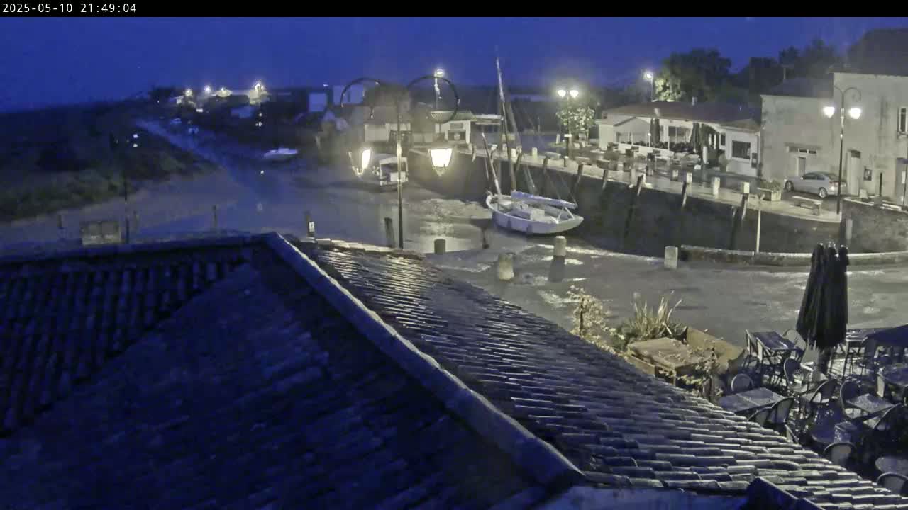 A nighttime view of a small harbor with boats, buildings, and outdoor seating, under a dark, possibly rainy sky.