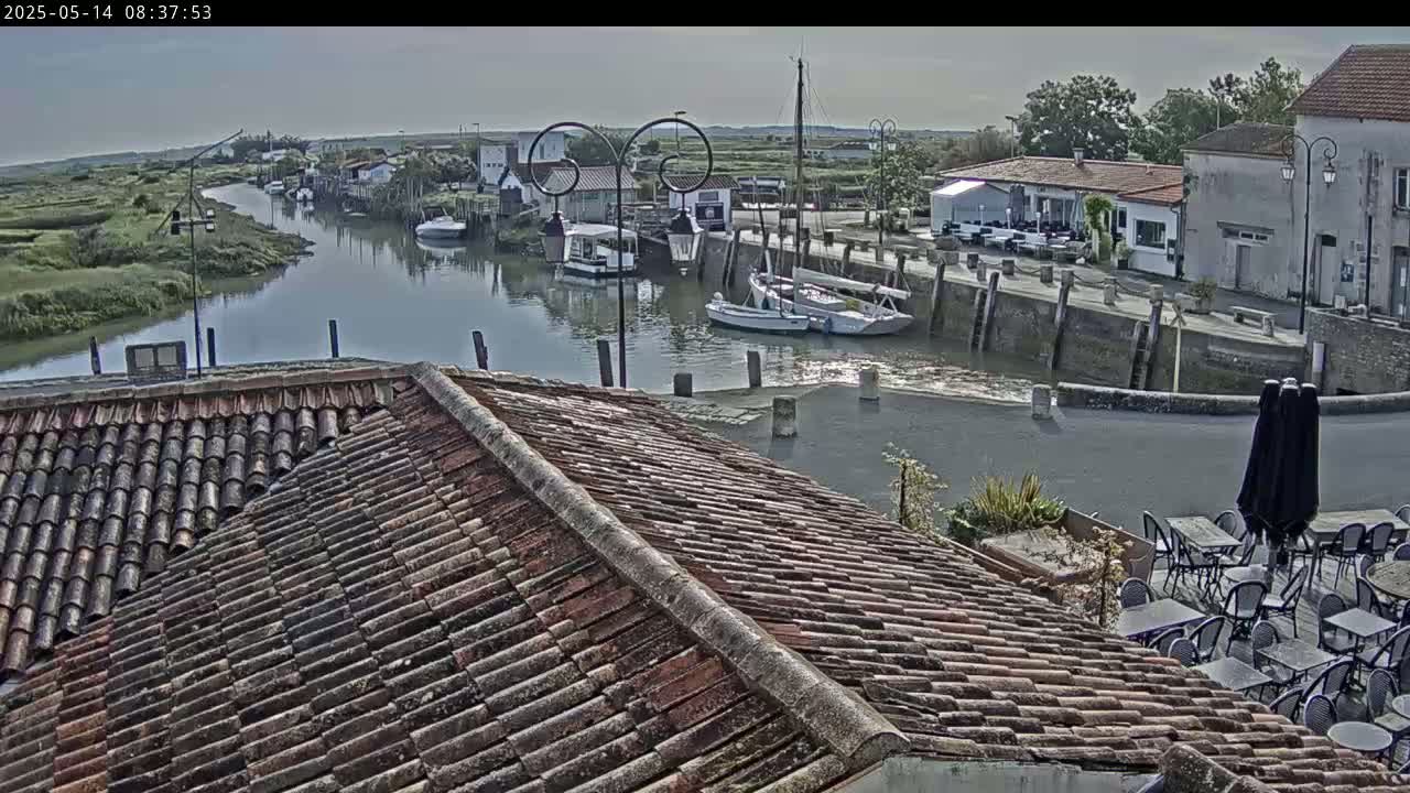 A tiled roof overlooks a calm waterway with boats docked near buildings in a village under a mostly cloudy sky.