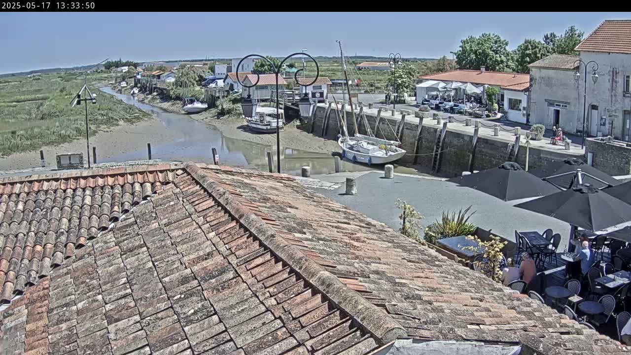 A tiled roof overlooks a harbor scene on a sunny day, with boats docked and people seated at outdoor tables near waterfront buildings.
