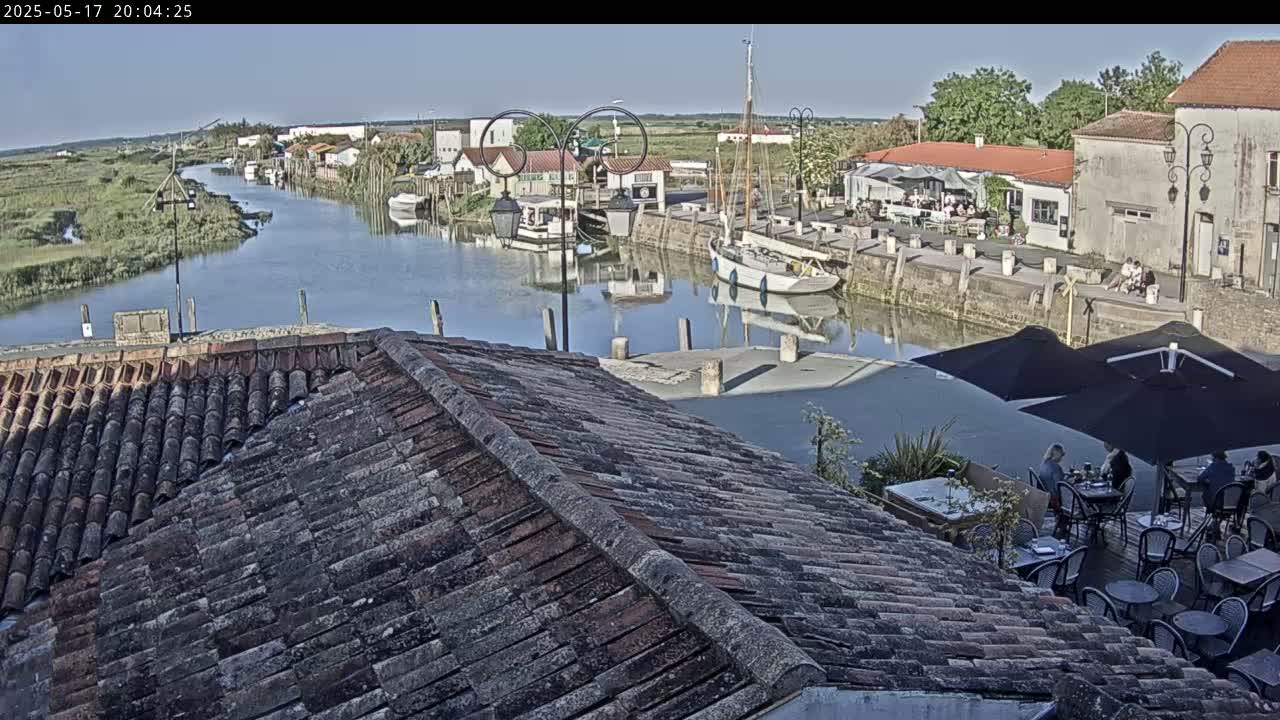 Overlooking a calm canal in a sunny village, boats are moored alongside buildings with outdoor seating areas.